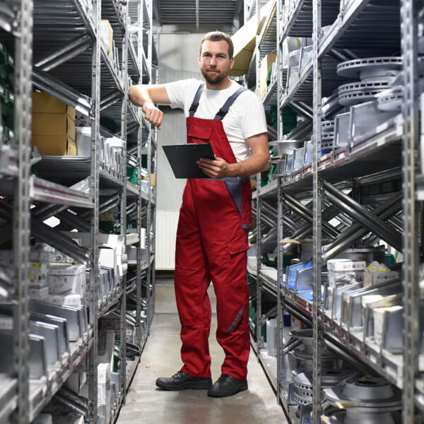 Technician in red overalls holding clipboard in auto parts warehouse.