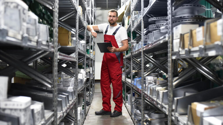 Technician in red overalls holding clipboard in auto parts warehouse.