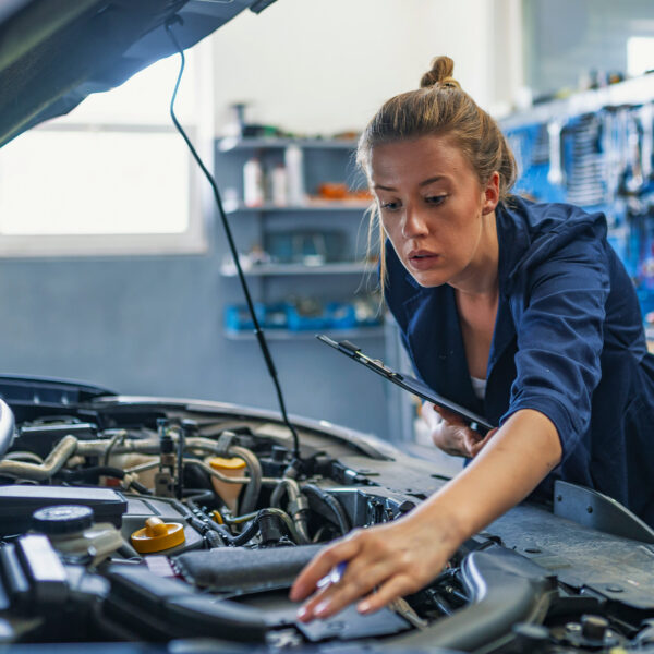 Female technician with clipboard examining a car engine.