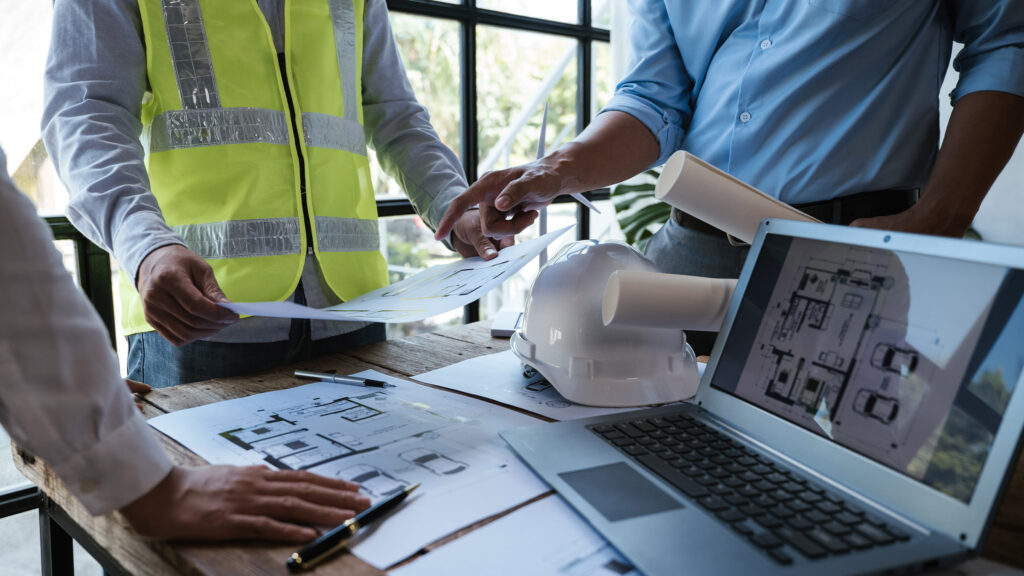 People wearing safety vests reviewing construction drawings and project plans on a table with a laptop and hard hat during a building design meeting.
