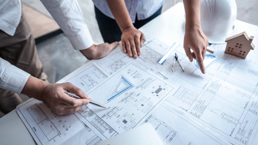 Hands of construction professionals reviewing architectural drawings and plans on a table with safety equipment nearby.