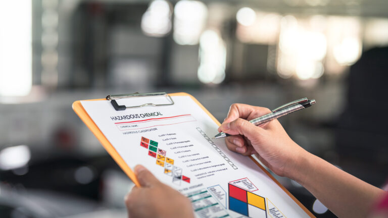 Person holding a clipboard and pen, reviewing a hazardous chemical form with safety symbols.
