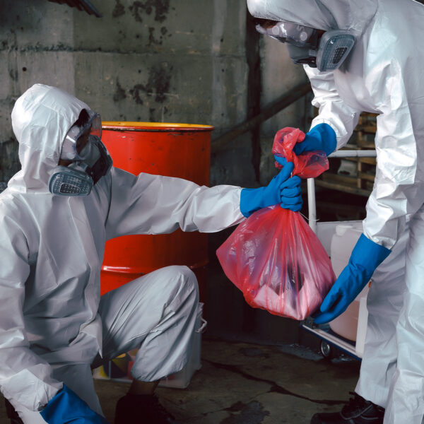 Two people wearing full protective suits and gloves handling a red waste bag near chemical drums.
