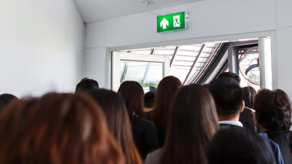 Group of people exiting a building through a door under a green emergency exit sign.