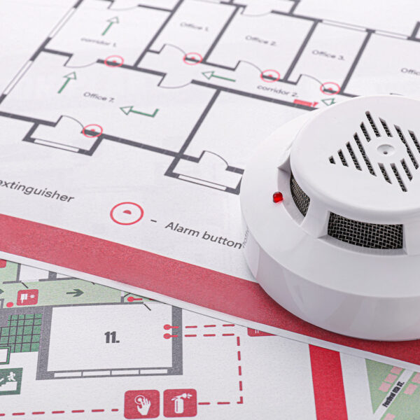 Smoke detector resting on a printed fire evacuation plan marked with exits and safety icons.