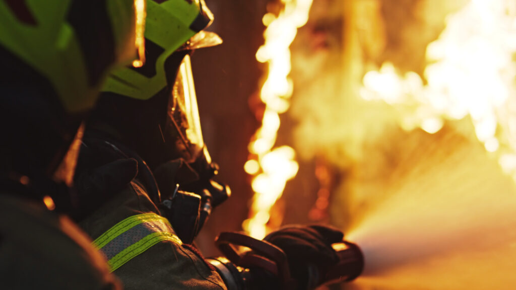 Firefighter in protective gear spraying water on flames.