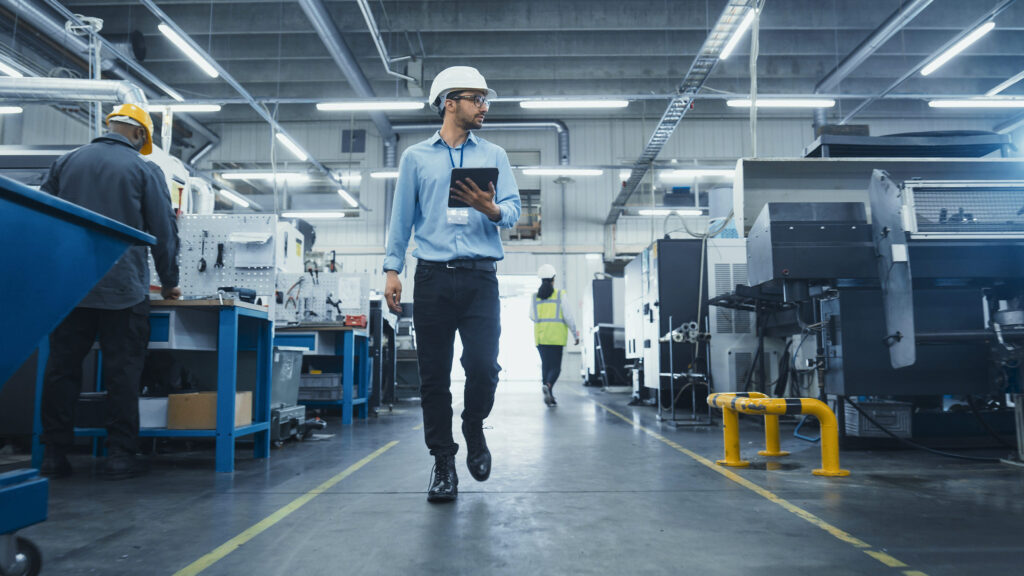 Factory manager wearing PPE and holding a tablet while walking through an engineering workshop.