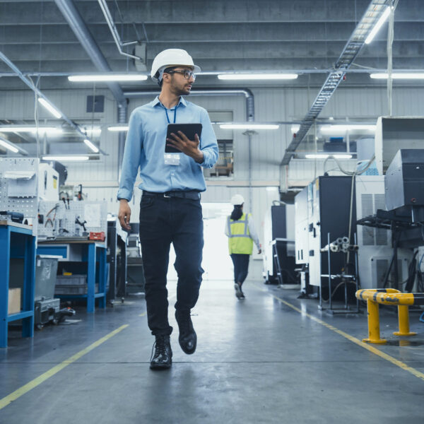 Factory manager wearing PPE and holding a tablet while walking through an engineering workshop.
