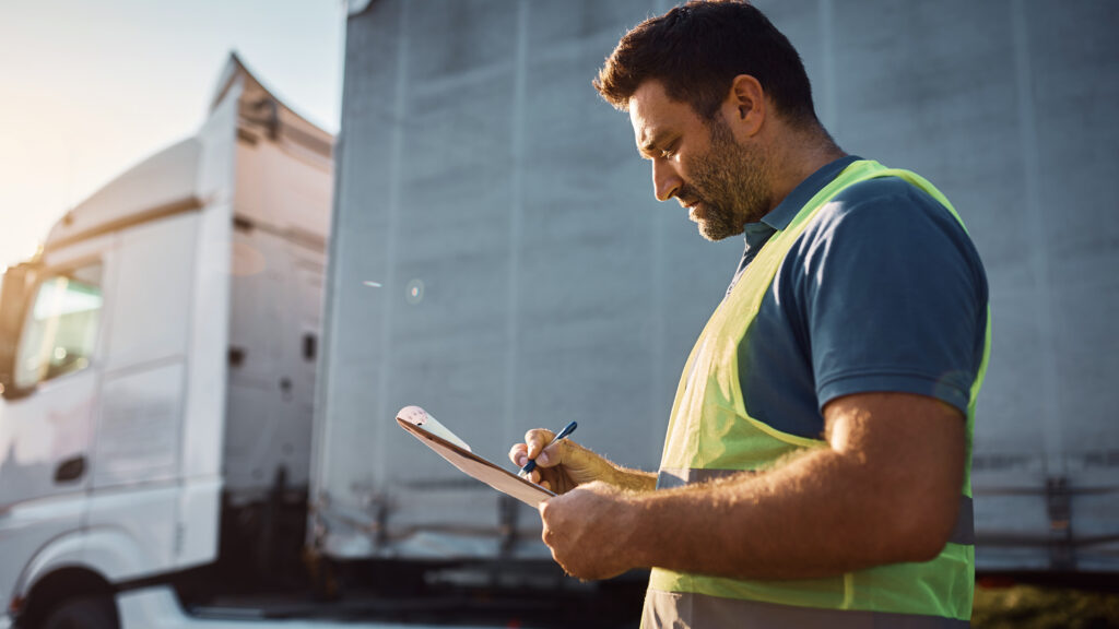 HGV driver wearing a safety vest reviewing delivery paperwork beside a lorry.