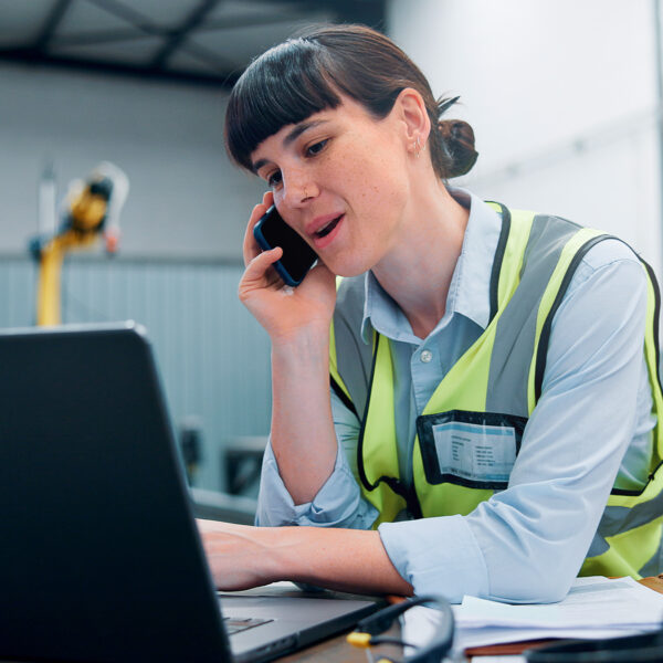 Warehouse supervisor wearing a high-vis vest talking on the phone and using a laptop.