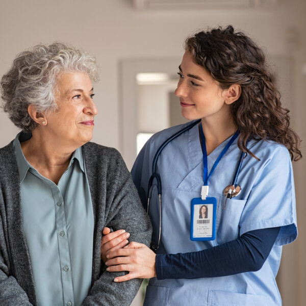 Nurse helping an older woman walk through a care home corridor.