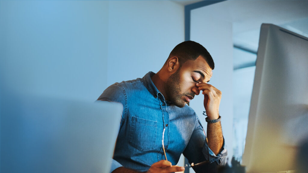 Office worker sitting at a desk, holding glasses and rubbing eyes while looking stressed or tired in front of computer screens.