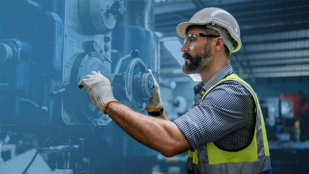 Factory worker wearing a hard hat, safety glasses and gloves, using industrial machinery in a manufacturing environment.