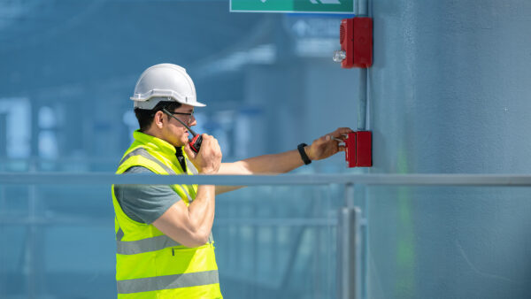 Person wearing a hard hat and safety vest speaking into a radio while pressing a red emergency alarm on a wall.