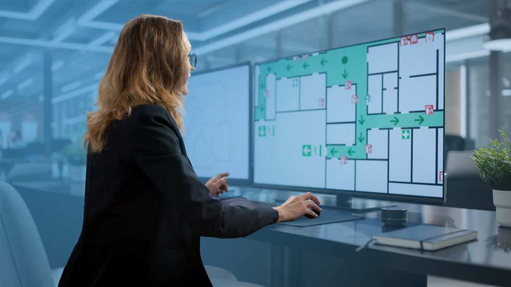 Office worker reviewing a digital building floor plan with evacuation routes displayed on multiple computer screens.