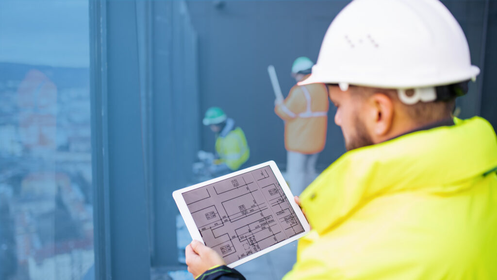Construction worker wearing a hard hat and high-visability jacket reviewing building plans on a tablet while colleagues work in the background.