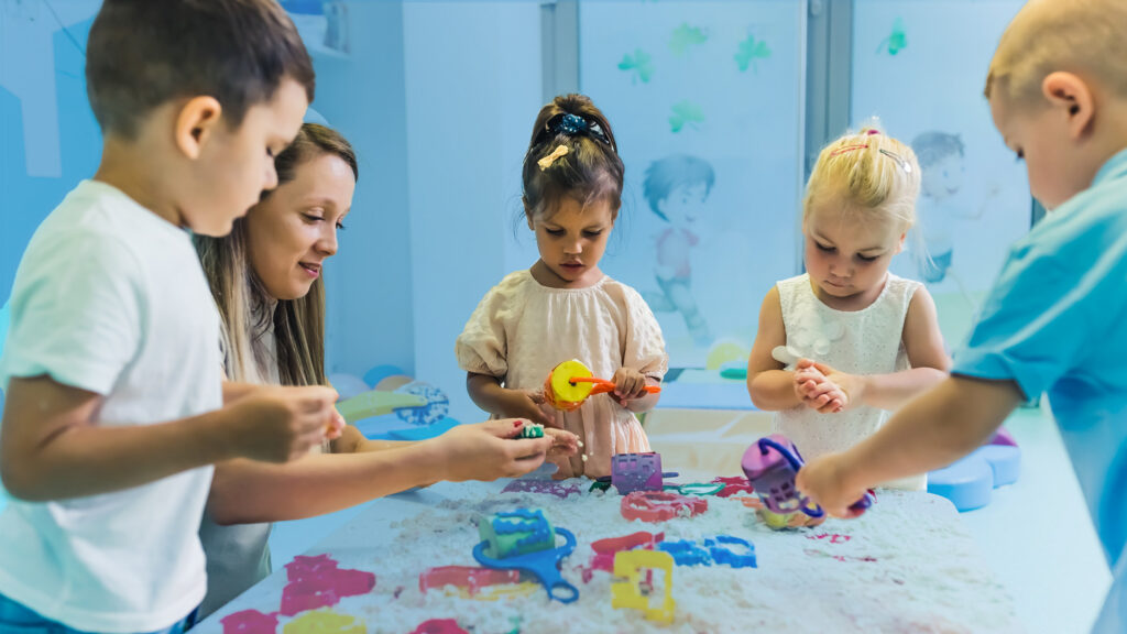 A group of young children are playing with colourful toys in a sensory sand table, supervised by an adult in a bright, child-friendly indoor classroom decorated with playful wall graphics.
