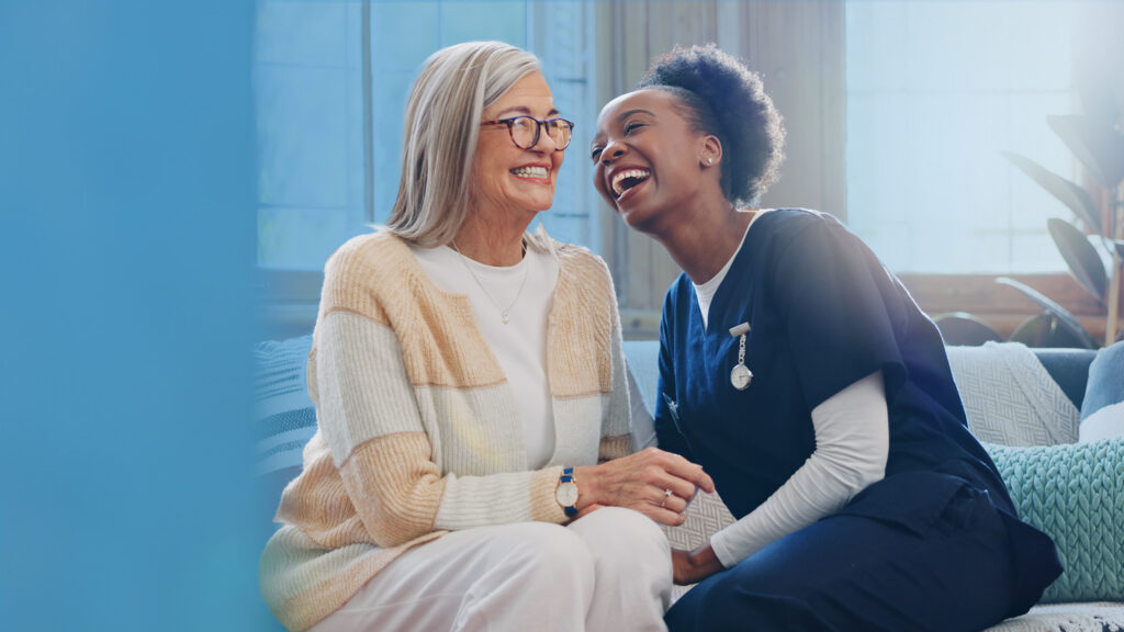 A smiling older woman sits on a sofa holding hands with a smiling care professional in navy scrubs; they are laughing together in a bright, homely living-room setting with a soft blue gradient on the left.