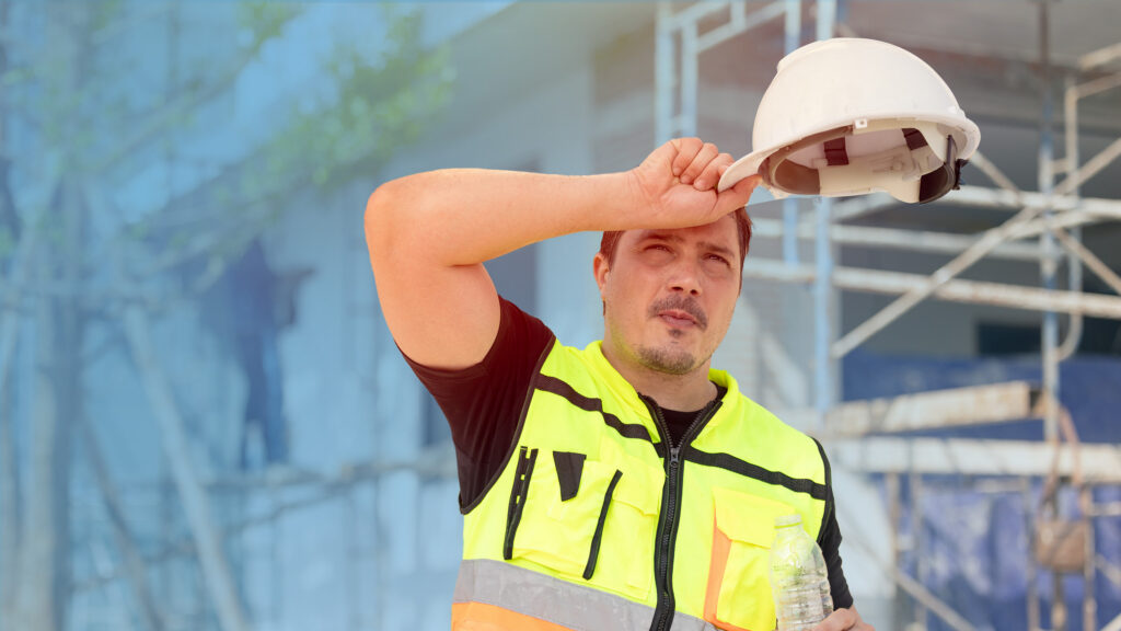 A construction worker in a high-visibility vest wipes sweat from his forehead while holding a hard hat and a water bottle, standing in front of scaffolding on a sunny day.