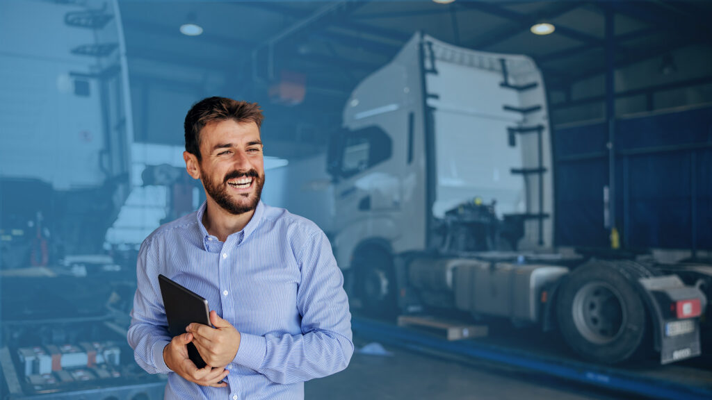 Smiling man in a blue shirt holding a tablet, standing in a lorry workshop with a large truck cab and equipment blurred in the background.