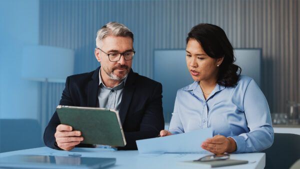 Two colleagues sitting at a desk in a modern office, reviewing documents together on a tablet and printed papers.