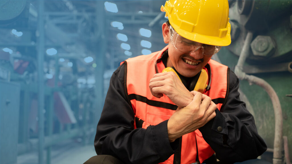 Factory worker in high‑vis vest and hard hat kneeling by machinery, clutching his painful wrist with a pained expression in an industrial workshop.
