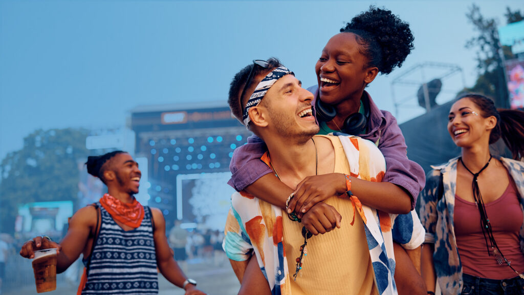 Young friends laughing and walking through an outdoor music festival, with one woman on a man’s back, colourful outfits, and a lit stage and crowd in the background.