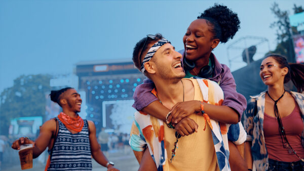 Young friends laughing and walking through an outdoor music festival, with one woman on a man’s back, colourful outfits, and a lit stage and crowd in the background.