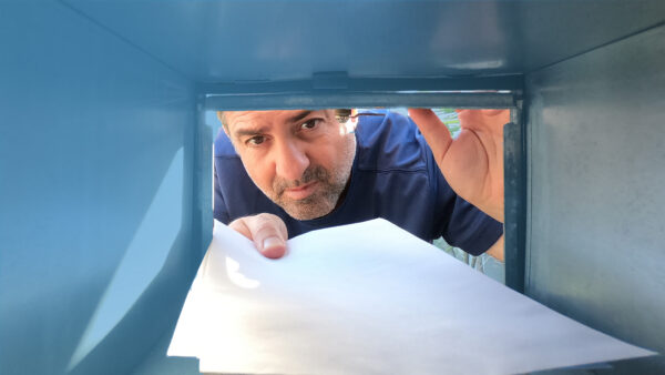 A man is peering into what looks like the inside of a metal letterbox, holding the flap open with one hand while inserting or retrieving a white envelope.