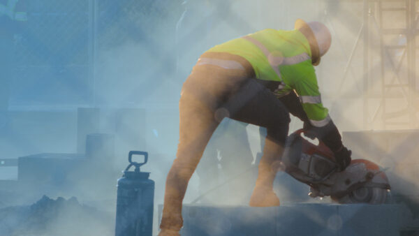 A construction worker wearing a high‑visibility jacket and hard hat is cutting concrete blocks with a power saw on a dusty building site, seen through a wire fence in warm, hazy light.